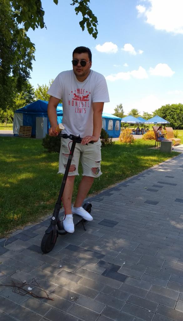 Mężczyzna w okularach przeciwsłonecznych i podartych szortach jedzie na hulajnodze elektrycznej po brukowanej ścieżce w parku, trzymając w ręku loda.