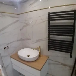 Wykończona łazienka z umywalką nablatową, złotą armaturą i grzejnikiem drabinkowym na tle marmurowych płytek z subtelnymi złotymi akcentami.