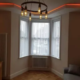 Jasne wnętrze pokoju z wykuszem, oświetleniem LED na suficie i drewnianą podłogą w jodełkę. Widoczny grzejnik i minimalistyczny żyrandol.