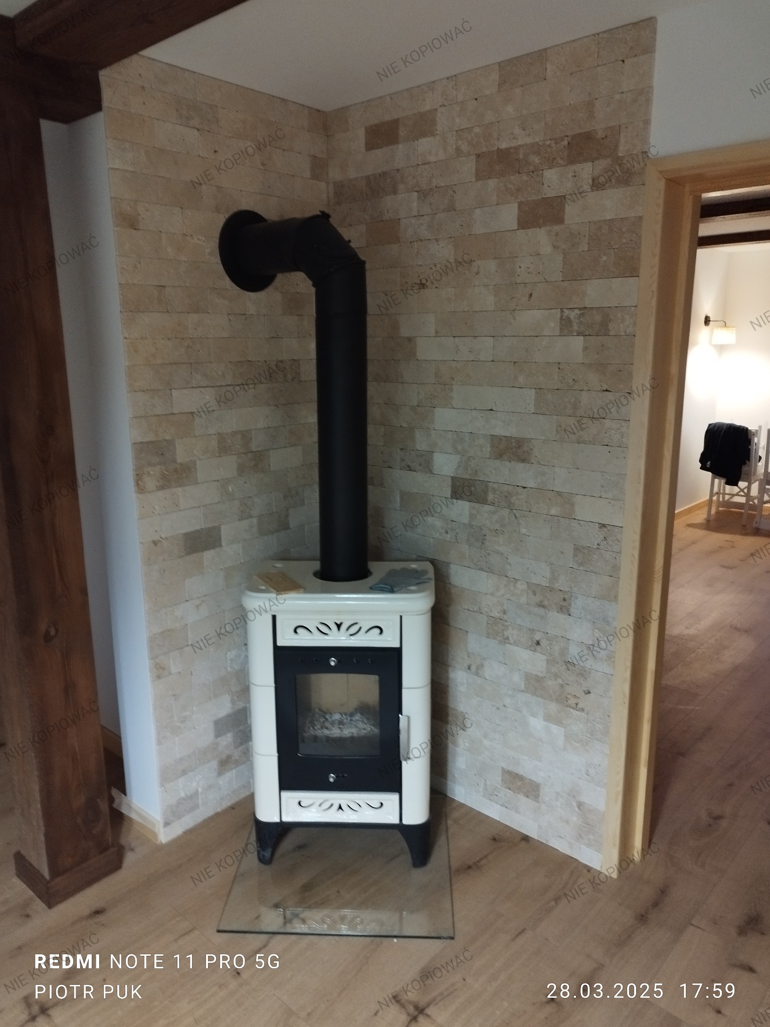 Biały piec wolnostojący z czarną rurą odprowadzającą spaliny, ustawiony w rogu pomieszczenia z wykończonymi ścianami z dekoracyjnego kamienia w odcieniach beżu, na podłodze panele imitujące drewno,...