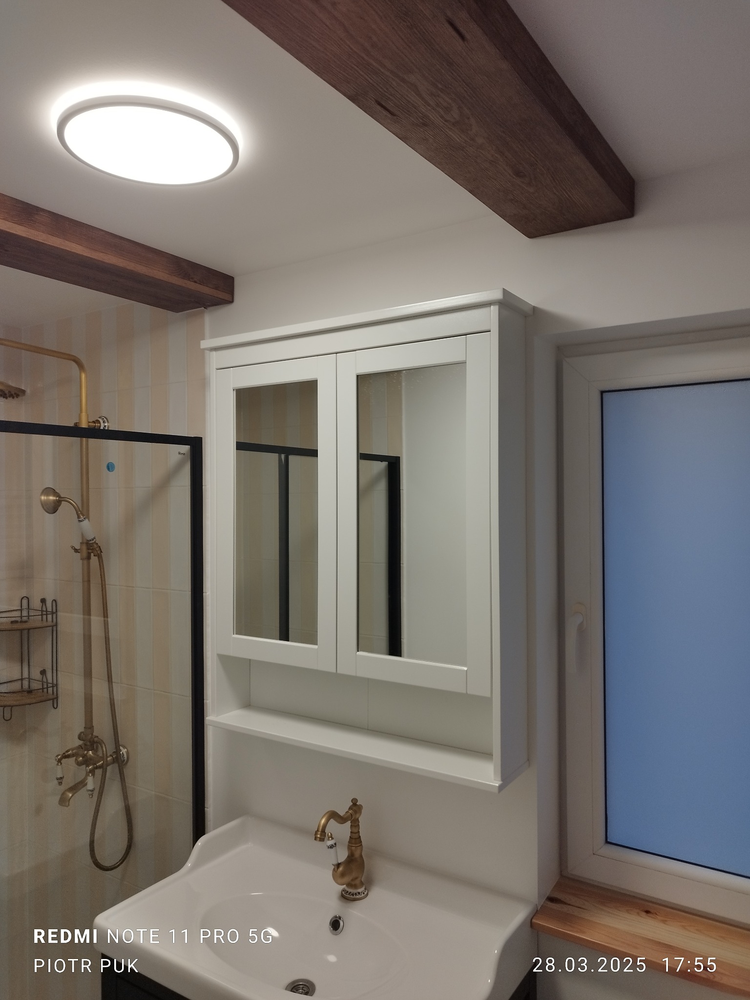 Wykończona łazienka z kabiną prysznicową w stylu retro z mosiężną armaturą i białą szafką z lustrami nad umywalką, drewniane belki na suficie.