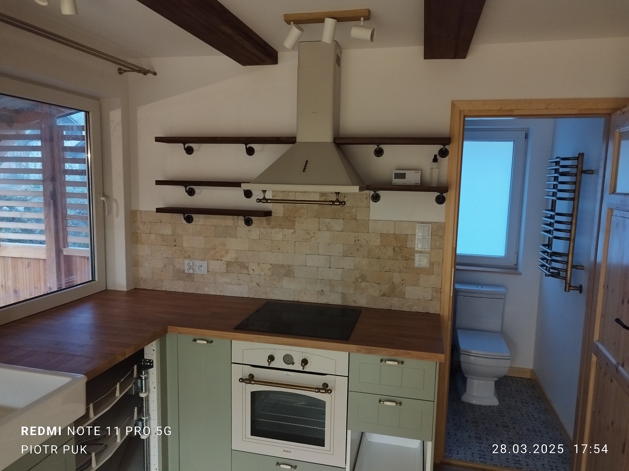 Nowoczesna kuchnia z elementami rustykalnymi: jasne, pistacjowe szafki, drewniany blat, kamienna mozaika na ścianie, półki z metalowymi wspornikami, widoczny fragment łazienki z toaletą...