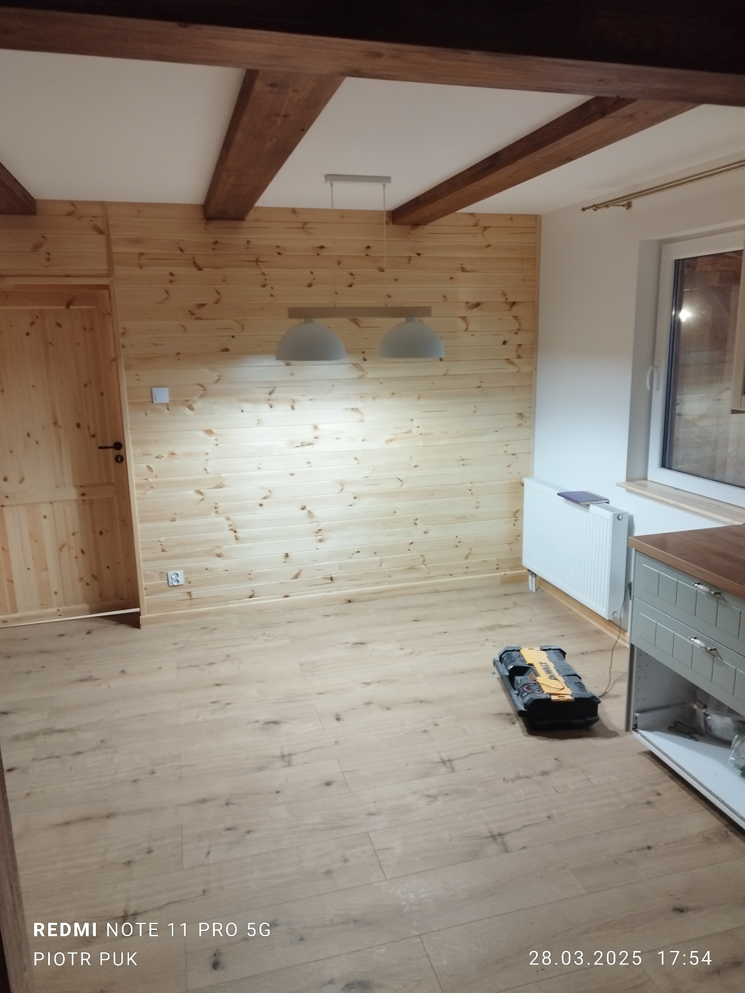 Wnętrze pokoju wykończonego boazerią z jasnego drewna, z widocznymi drewnianymi belkami na suficie, wiszącą lampą, grzejnikiem i skrzynką narzędziową na podłodze.