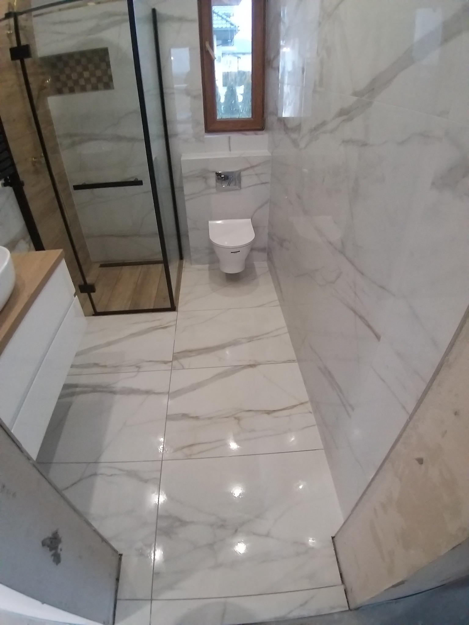 Wykończona łazienka z kabiną prysznicową typu walk-in, białą armaturą i płytkami imitującymi marmur na ścianach i podłodze. Widok na okno i fragment szafki z umywalką.