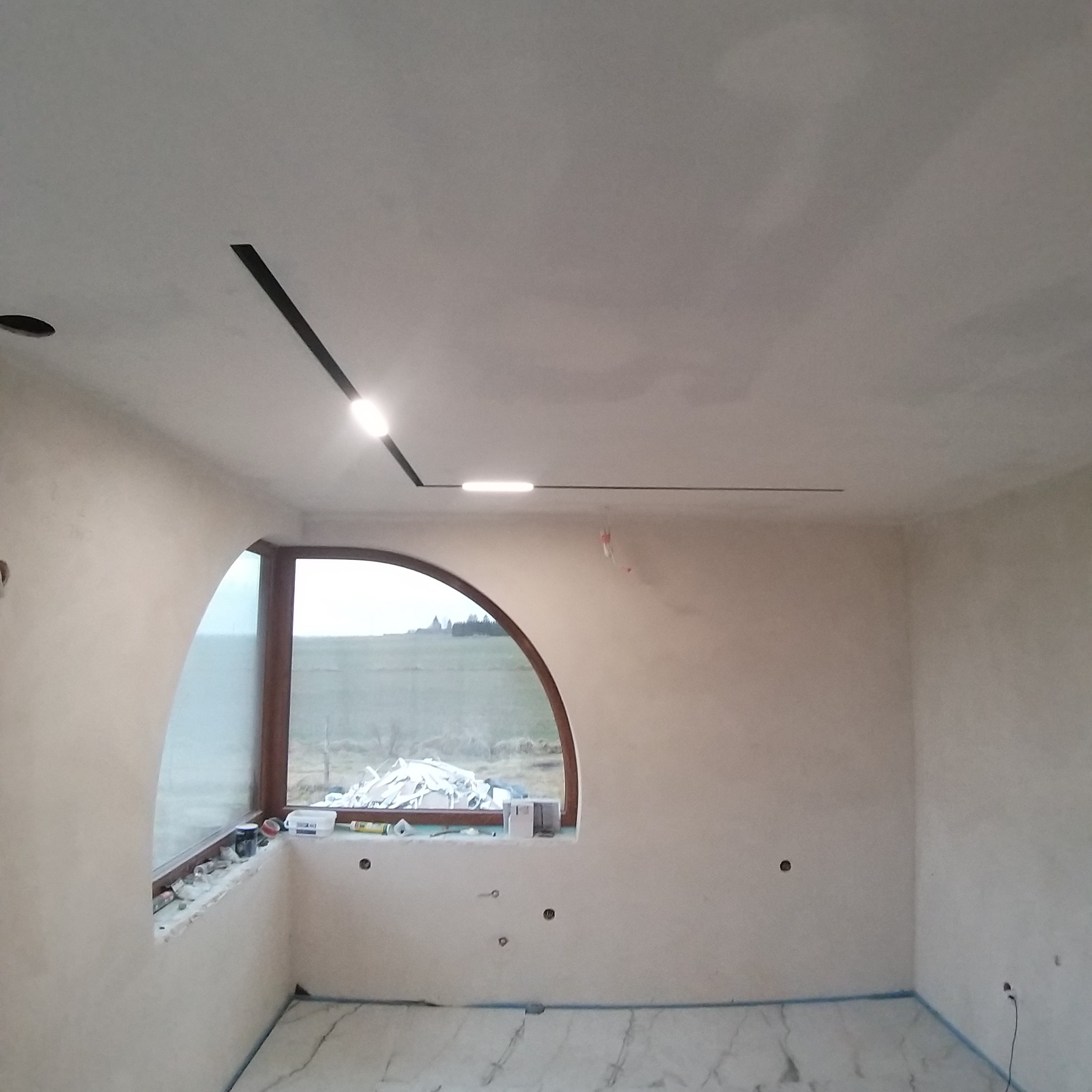 Ściany po szpachlowaniu w Zamościu, widoczne listwy oświetleniowe na suficie i okno o nietypowym kształcie. Przygotowanie do dalszych prac wykończeniowych.