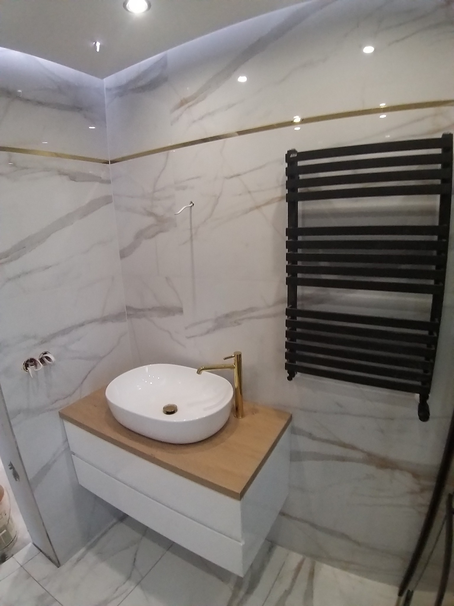 Wykończona łazienka z umywalką nablatową, złotą armaturą i grzejnikiem drabinkowym na tle marmurowych płytek z subtelnymi złotymi akcentami.