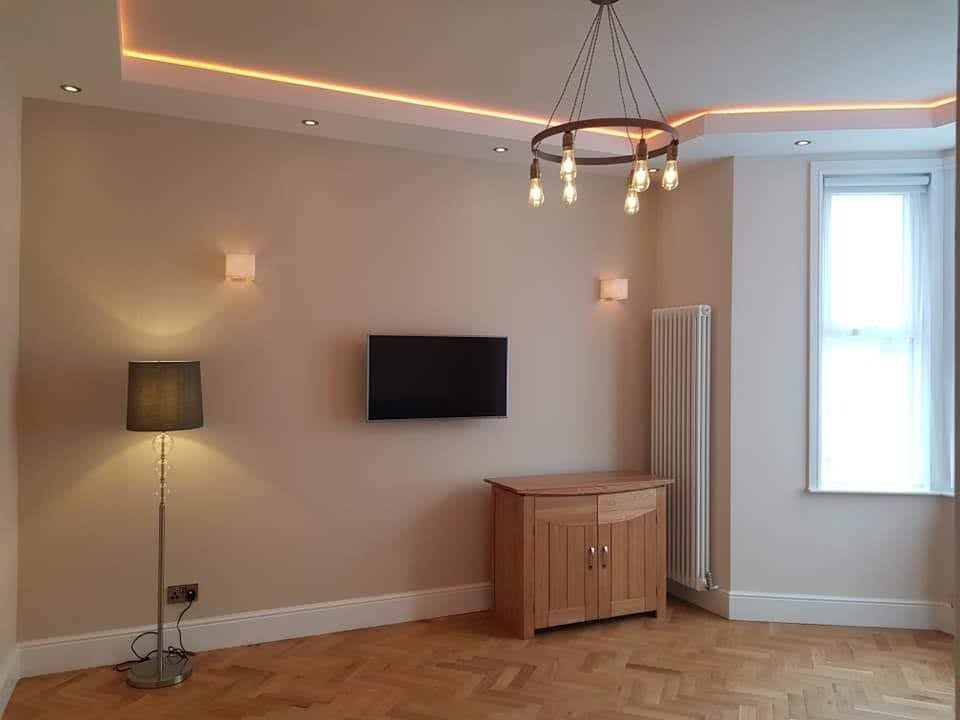 Jasne wnętrze po remoncie: parkiet w jodełkę, kremowe ściany, oświetlenie LED na suficie, wisząca lampa, szafka z drewna i telewizor na ścianie.