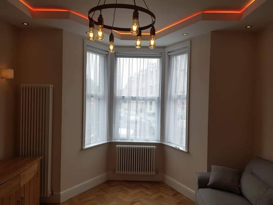 Jasne wnętrze pokoju z wykuszem, oświetleniem LED na suficie i drewnianą podłogą w jodełkę. Widoczny grzejnik i minimalistyczny żyrandol.