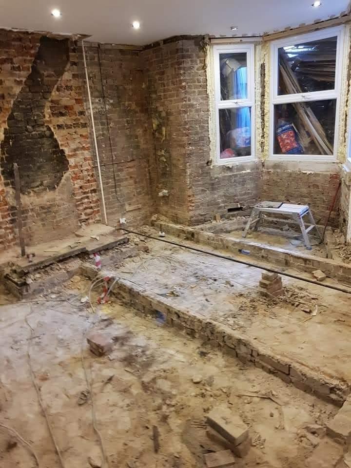 Wnętrze w trakcie remontu: odsłonięta cegła, okna z drewnianą stolarką i prowizoryczne rusztowanie. Widoczne ślady demontażu podłogi i instalacji elektrycznej.