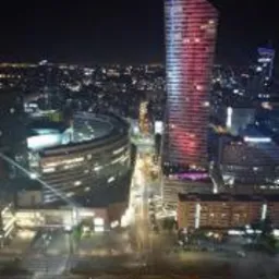 Nocny widok na panoramę Warszawy z wieżowcem Warsaw Spire oświetlonym na czerwono w centrum kadru, widoczne ulice i budynki w blasku świateł.