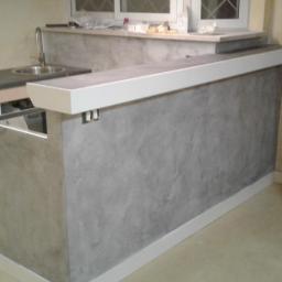 resinart - Surowy stan wykończenia baru kuchennego z szarą, teksturowaną ścianą i białymi listwami, widoczna instalacja zlewu i okno w tle.