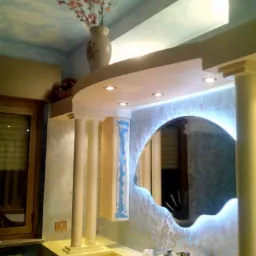 Oryginalna łazienka po remoncie z dekoracyjnym lustrem podświetlonym LED, kolumnami, i malowanym sufitem imitującym niebo, oraz wazonem z kwiatami na półce.