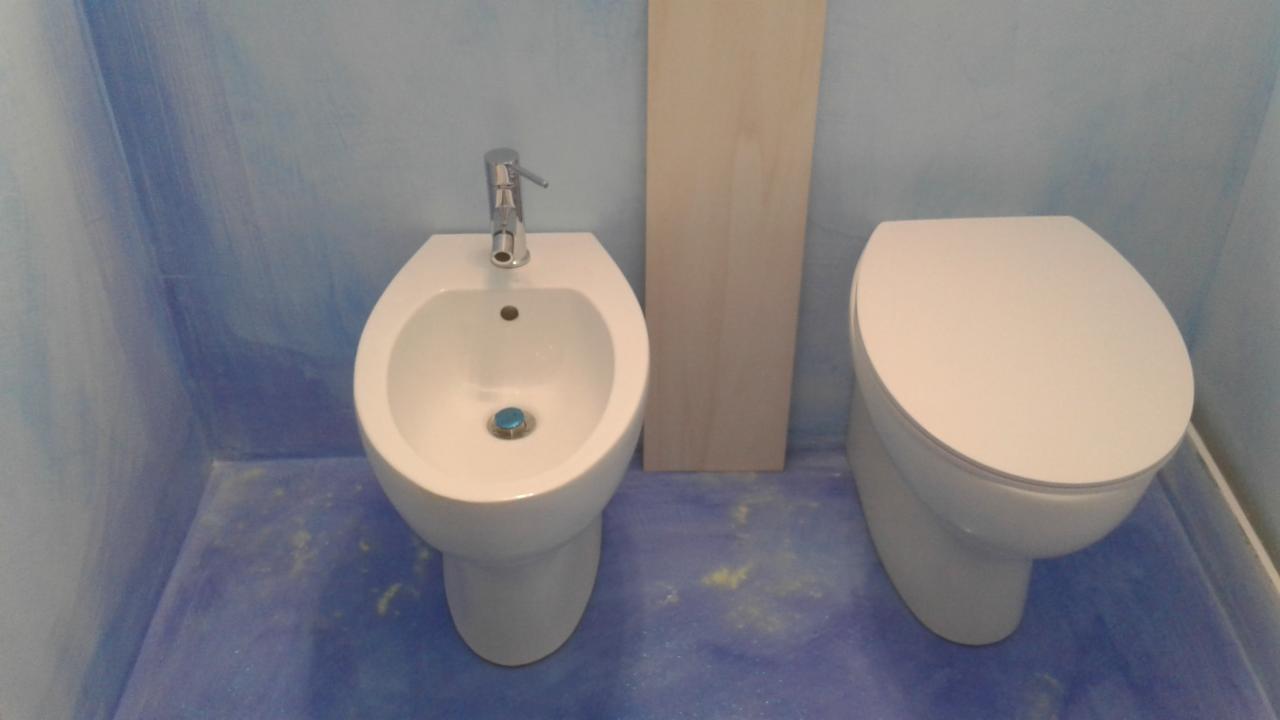 Nowoczesna łazienka z bidetem i toaletą, ściany w odcieniach błękitu z efektem malarskim, podłoga w kolorze niebieskim z żółtymi zabrudzeniami po remoncie, obok bidetu stoi drewniana deska.