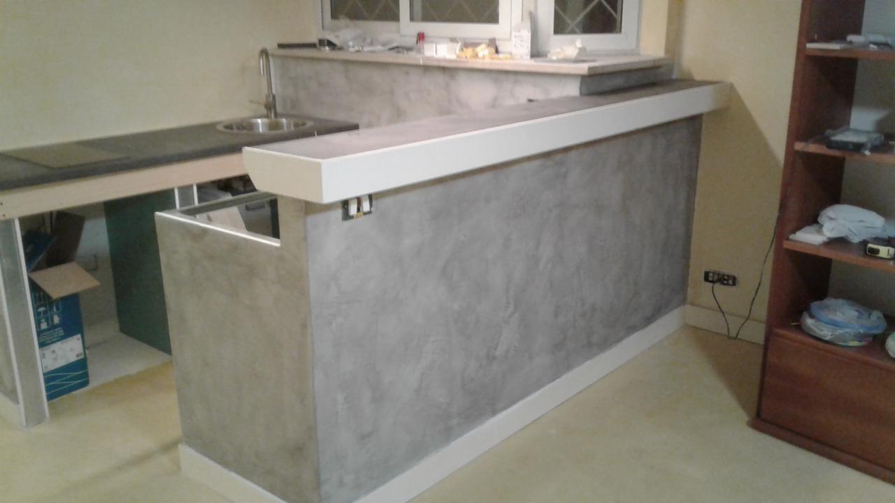 Surowy stan wykończenia baru kuchennego z szarą, teksturowaną ścianą i białymi listwami, widoczna instalacja zlewu i okno w tle.