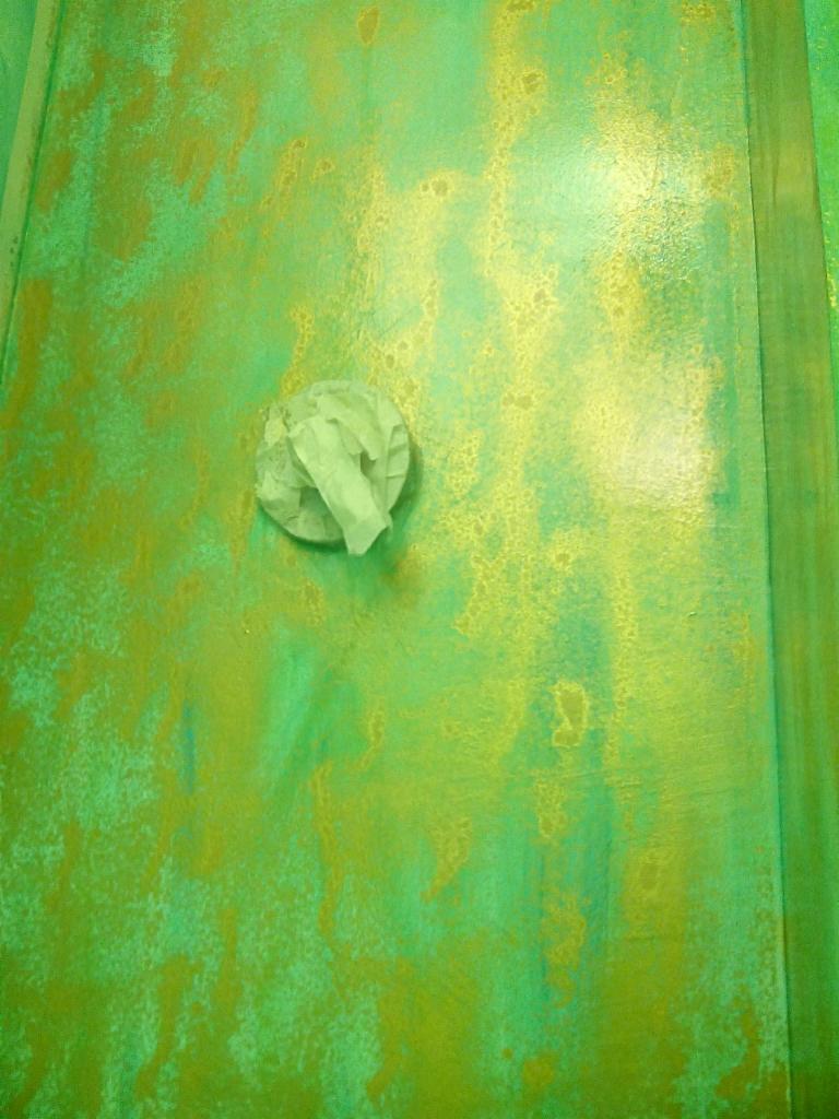 Ściana pokryta nieregularną, zielono-złotą fakturą, z przyklejoną kulką z papieru maskującego, prawdopodobnie w trakcie prac malarskich.