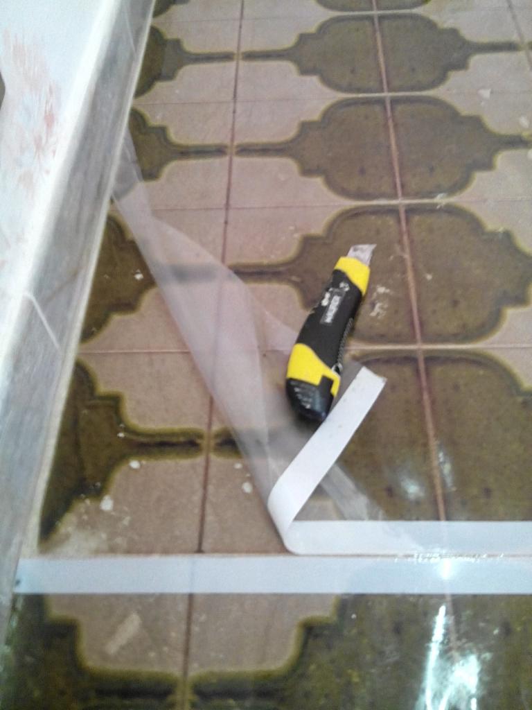 Niedokładne ujęcie fragmentu podłogi w łazience z wzorzystymi, brązowo-beżowymi płytkami, zabezpieczonej folią i taśmą malarską, obok leży nóż segmentowy z żółto-czarną rękojeścią.