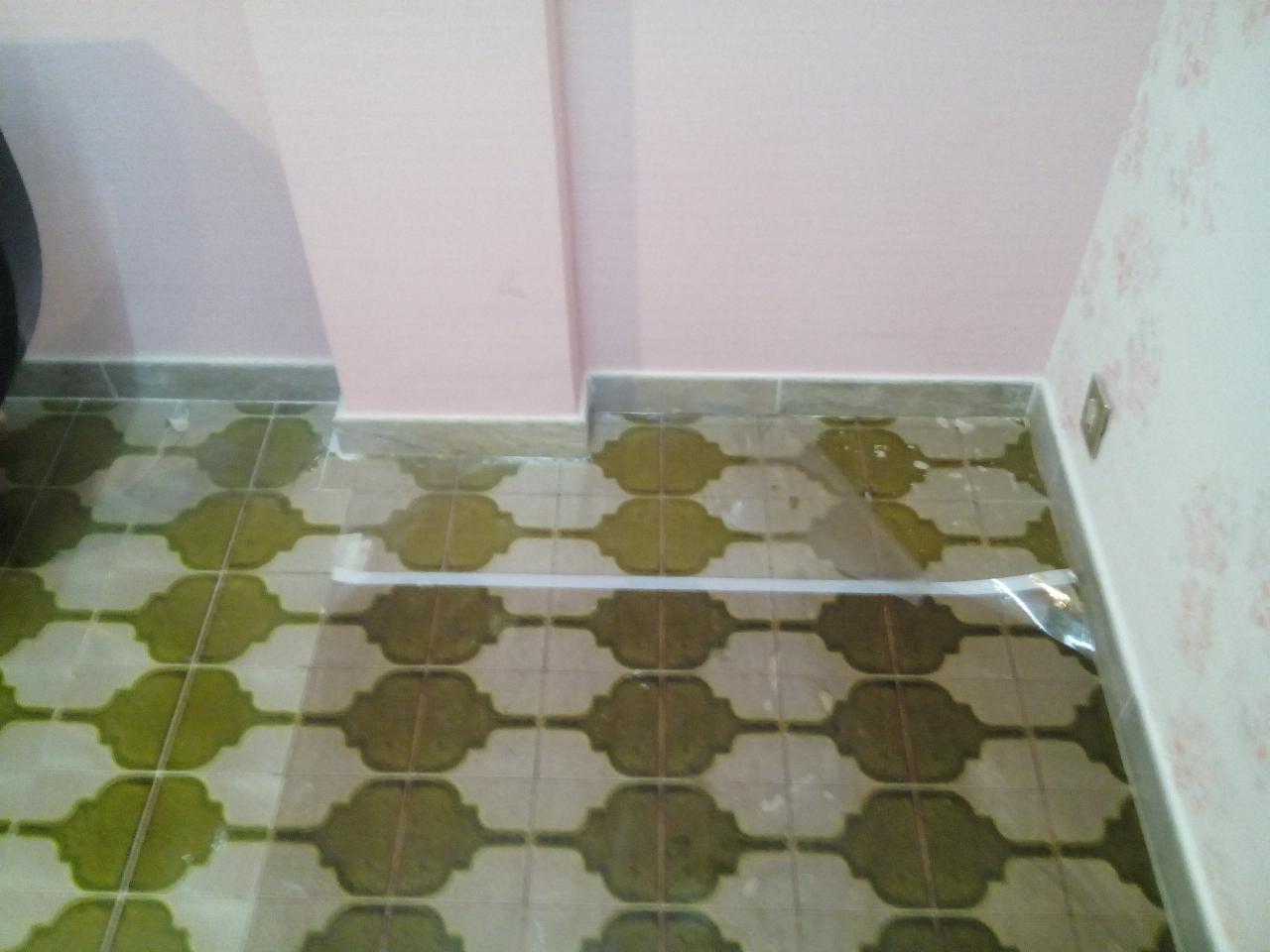 Fragment łazienki w trakcie prac: ułożona glazura podłogowa w odcieniach zieleni i beżu, zabezpieczona folią malarską, różowa ściana w górnej części, fragment ściany z tapetą w kwiaty po prawej.