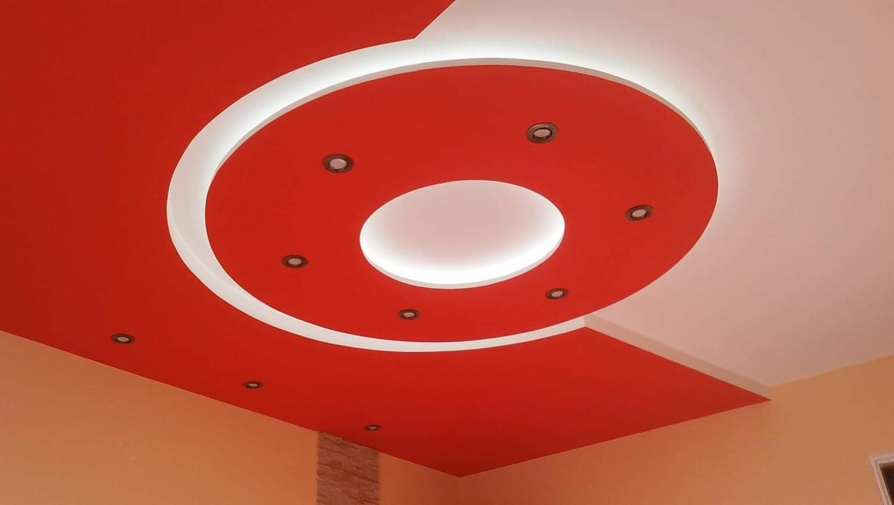 Czerwony sufit podwieszany z okrągłym wgłębieniem oświetlonym białym światłem i punktowymi lampami, kontrastujący z białym fragmentem sufitu.