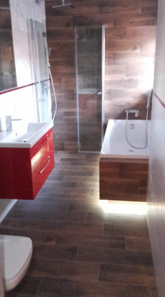 Nowoczesna łazienka z drewnopodobnymi płytkami, czerwona szafka pod umywalką, kabina prysznicowa i wanna z podświetleniem LED.