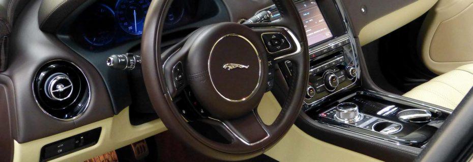 Wnętrze luksusowego samochodu Jaguar z brązową skórzaną kierownicą z chromowanym logo, beżową tapicerką i błyszczącą konsolą środkową