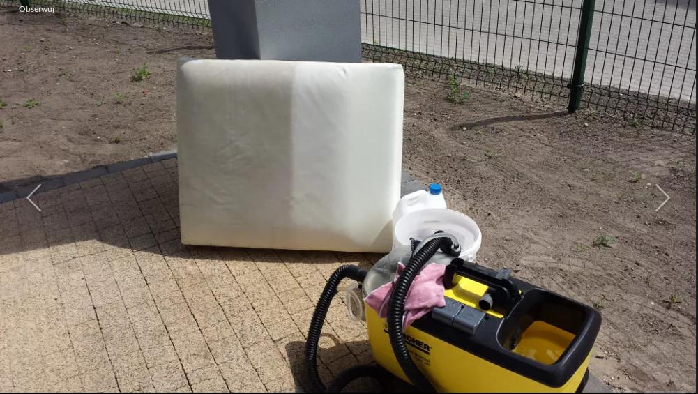 Kwadratowa poduszka tapicerowana, w połowie wyczyszczona, obok żółty odkurzacz piorący Karcher, wiadro i pojemnik z płynem na kostce brukowej.