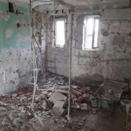 Wnętrze pomieszczenia w trakcie wyburzania: gruz, cegły, odsłonięte instalacje, fragment ściany z resztkami farby, okna w tle, taczka z resztkami materiałów.