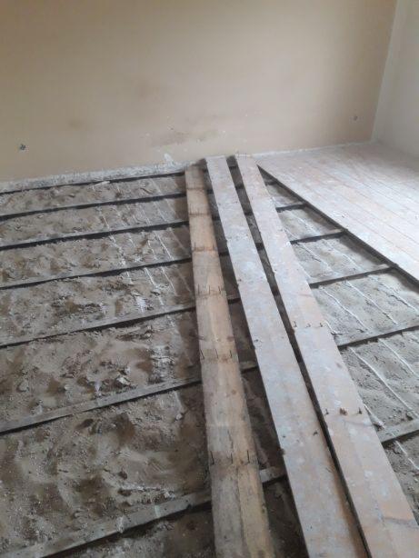 Demontaż starej podłogi: odsłonięte legary, fragmenty desek podłogowych i ślady zaprawy na podłożu w remontowanym pokoju.