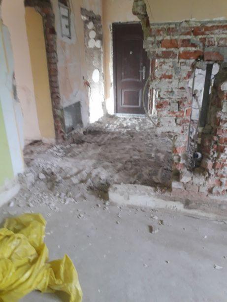 Wnętrze w trakcie demontażu: fragment zburzonej ściany z czerwonej cegły, odsłonięte przewody, gruz na podłodze, widoczna brązowa futryna drzwi, żółta torba na gruz w rogu.