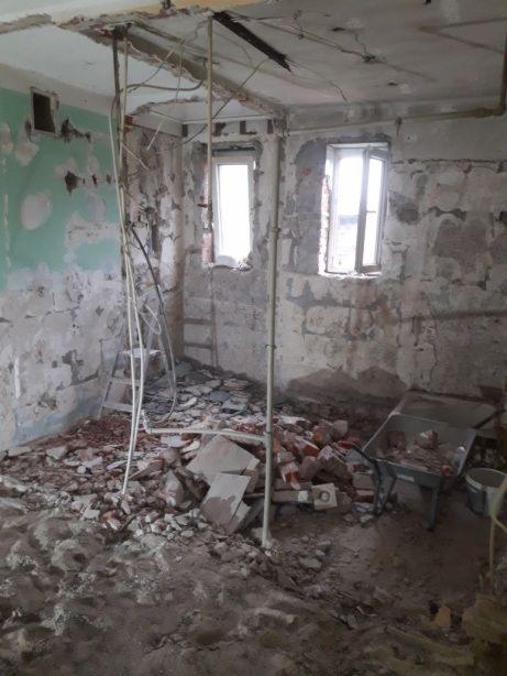 Wnętrze pomieszczenia w trakcie wyburzania: gruz, cegły, odsłonięte instalacje, fragment ściany z resztkami farby, okna w tle, taczka z resztkami materiałów.