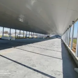 Wnętrze hali w trakcie budowy z betonową posadzką, stalowymi słupami i widocznymi workami z materiałami budowlanymi na zewnątrz, w tle budynki gospodarcze i pole.