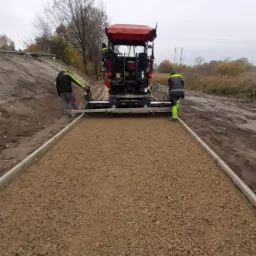 Układanie nawierzchni żwirowej za pomocą specjalistycznej maszyny z udziałem dwóch pracowników w odblaskowych strojach. Widoczny świeżo ułożony żwir ograniczony krawężnikami.
