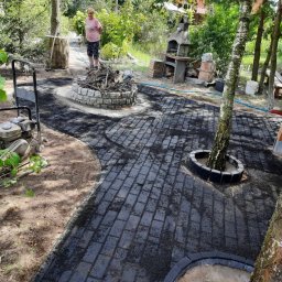 BRUK-COMPANY USŁUGI BRUKARSKIE NATALIA KOWALSKA - Ścieżka z kostki brukowej w ogrodzie, z obudową drzewa i paleniskiem. W tle kobieta i grill. Widoczna ubijarka do gruntu. Kostka świeżo ułożona, posypana piaskiem.
