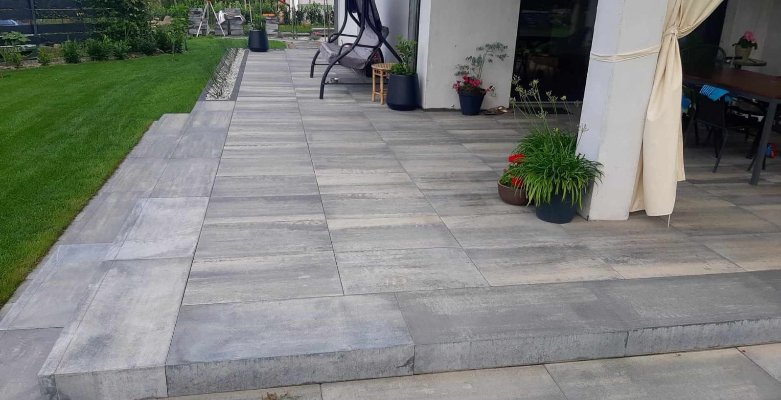 Nowoczesny taras z szarych płyt betonowych, z zielonym trawnikiem w tle i elementami wyposażenia: huśtawka, stolik, donice z kwiatami. Estetyczne wykończenie.