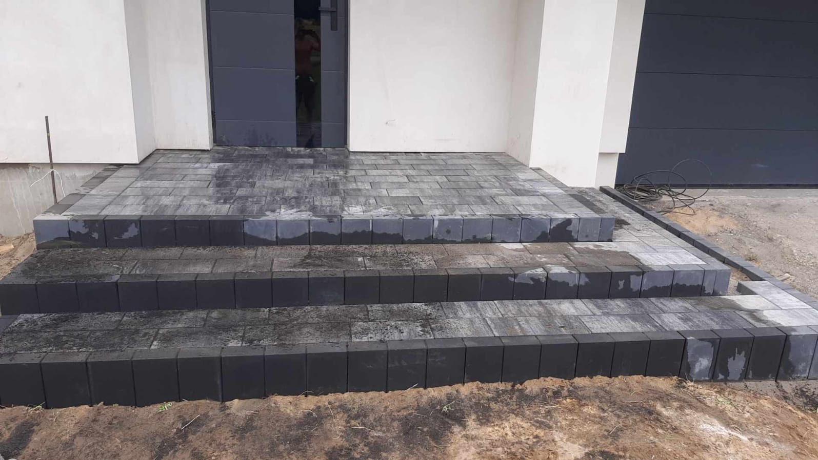 Nowe, trzystopniowe schody wejściowe z szarej kostki brukowej, prowadzące do ciemnych drzwi wejściowych. Widoczny fragment elewacji i brama garażowa.