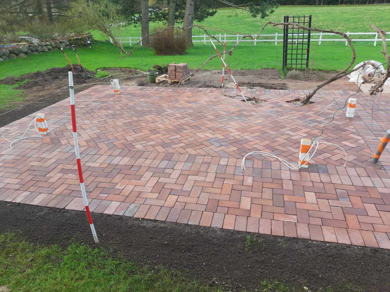 Układanie kostki brukowej w jodełkę na posesji, widoczne narzędzia pomiarowe i przygotowane podłoże. Częściowo ukończona nawierzchnia z kostki w odcieniach brązu i czerwieni.