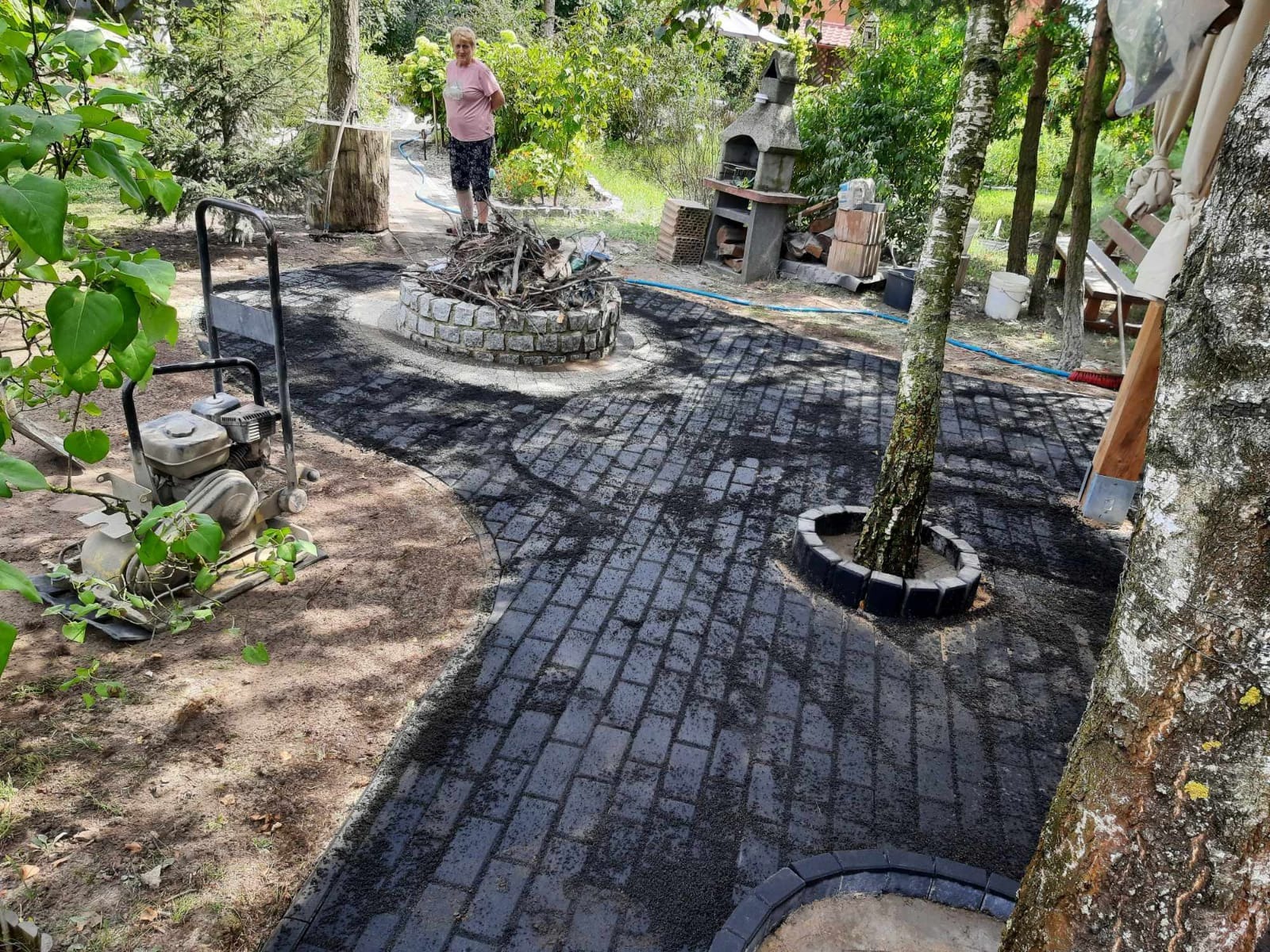 Ścieżka z kostki brukowej w ogrodzie, z obudową drzewa i paleniskiem. W tle kobieta i grill. Widoczna ubijarka do gruntu. Kostka świeżo ułożona, posypana piaskiem.