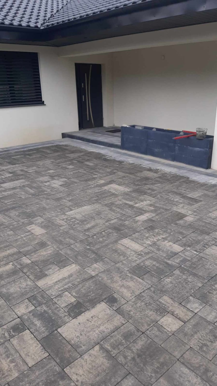 Ułożona szara kostka brukowa przed wejściem do domu z ciemnymi drzwiami i oknem z żaluzjami. Widoczny fragment dachu i donica z narzędziami.