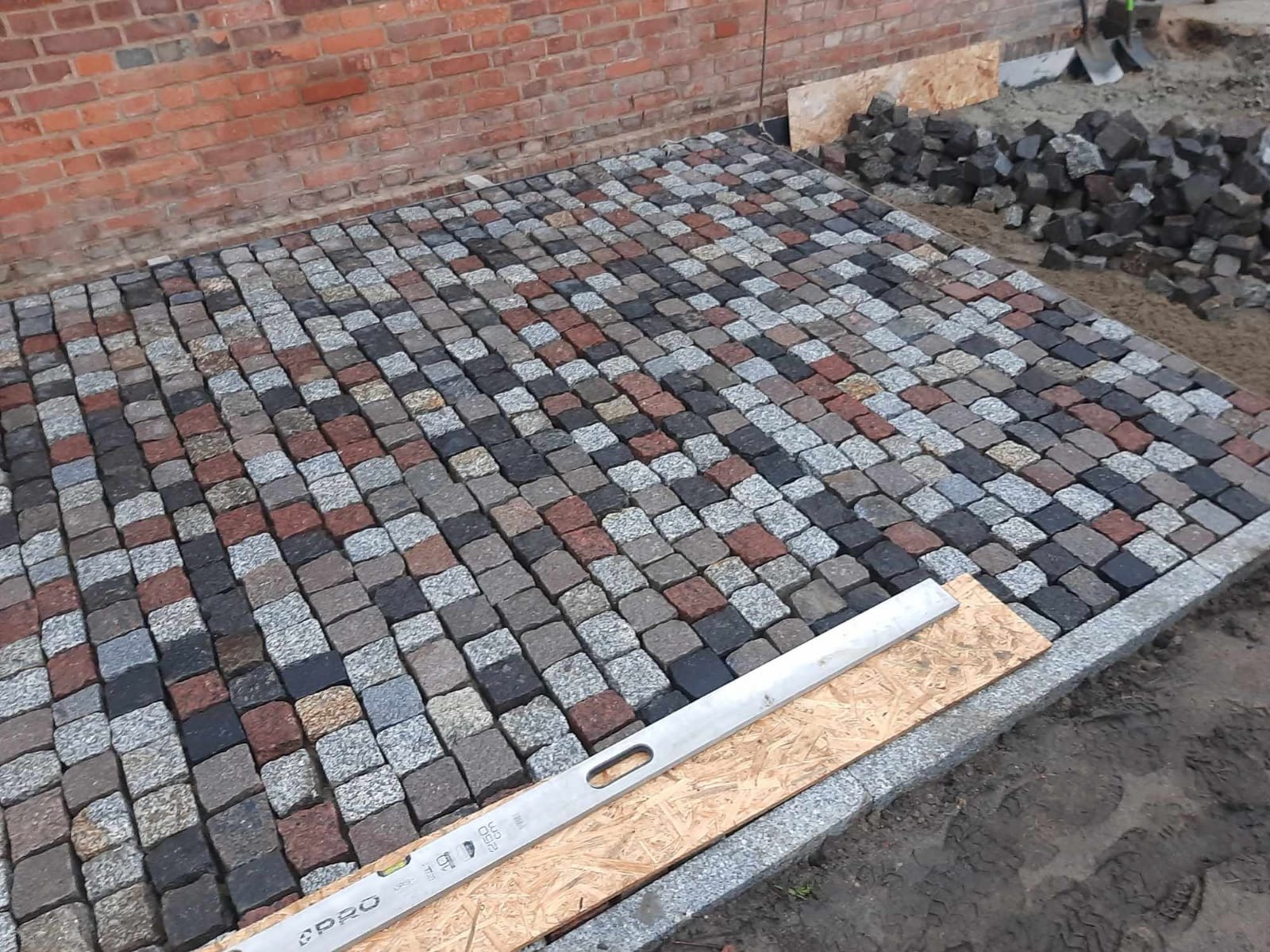Ułożona kostka brukowa w odcieniach szarości, brązu i czerni, z poziomicą na drewnianej płycie. W tle mur z czerwonej cegły i narzędzia.