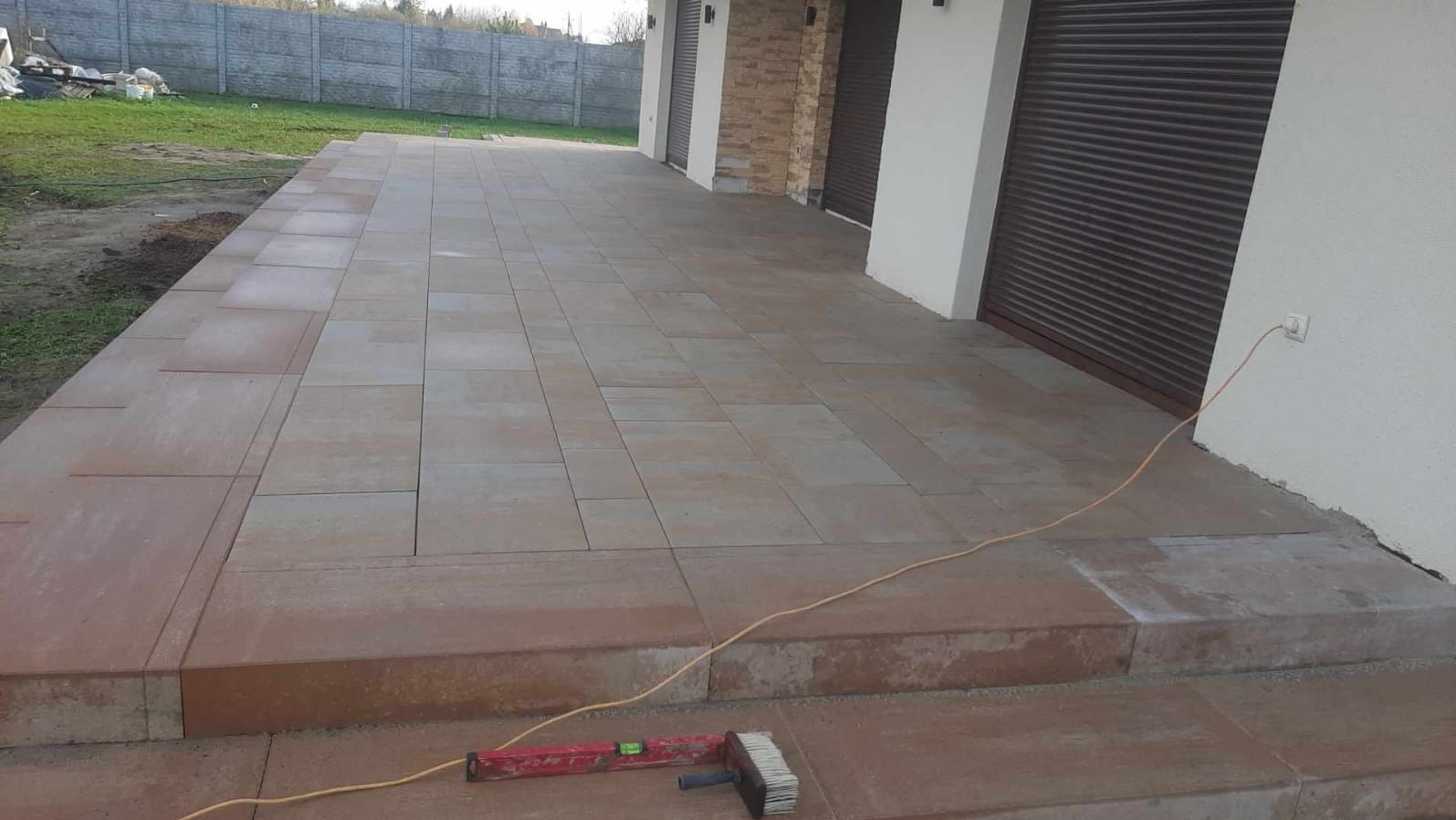 Ułożona kostka brukowa na tarasie domu z widocznymi narzędziami: poziomica i szczotka. Jasne, naturalne światło. Widok na budynek z roletami.