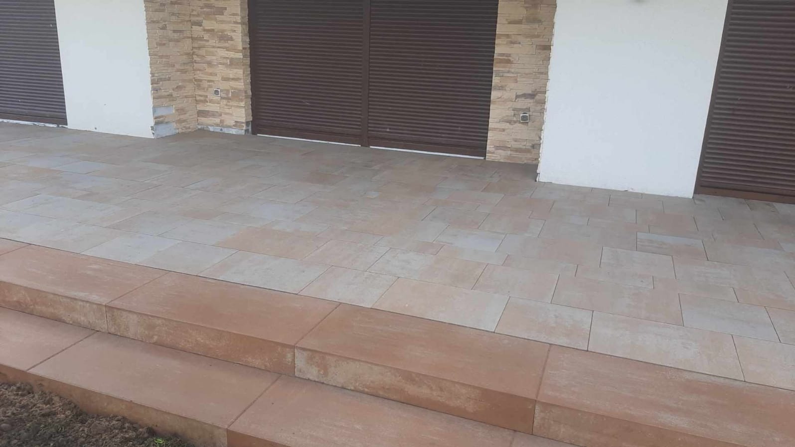 Ułożona kostka brukowa na tarasie z brązowymi schodami i zamkniętymi, brązowymi roletami w tle. Widoczne fragmenty elewacji z białym tynkiem i kamienną okładziną.