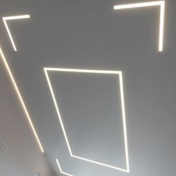 Firma Usługowa Majer - Biały sufit z wpuszczonymi liniowymi oprawami LED tworzącymi geometryczne wzory: kwadrat, kąty proste i linie. Widoczne fragmenty okna.
