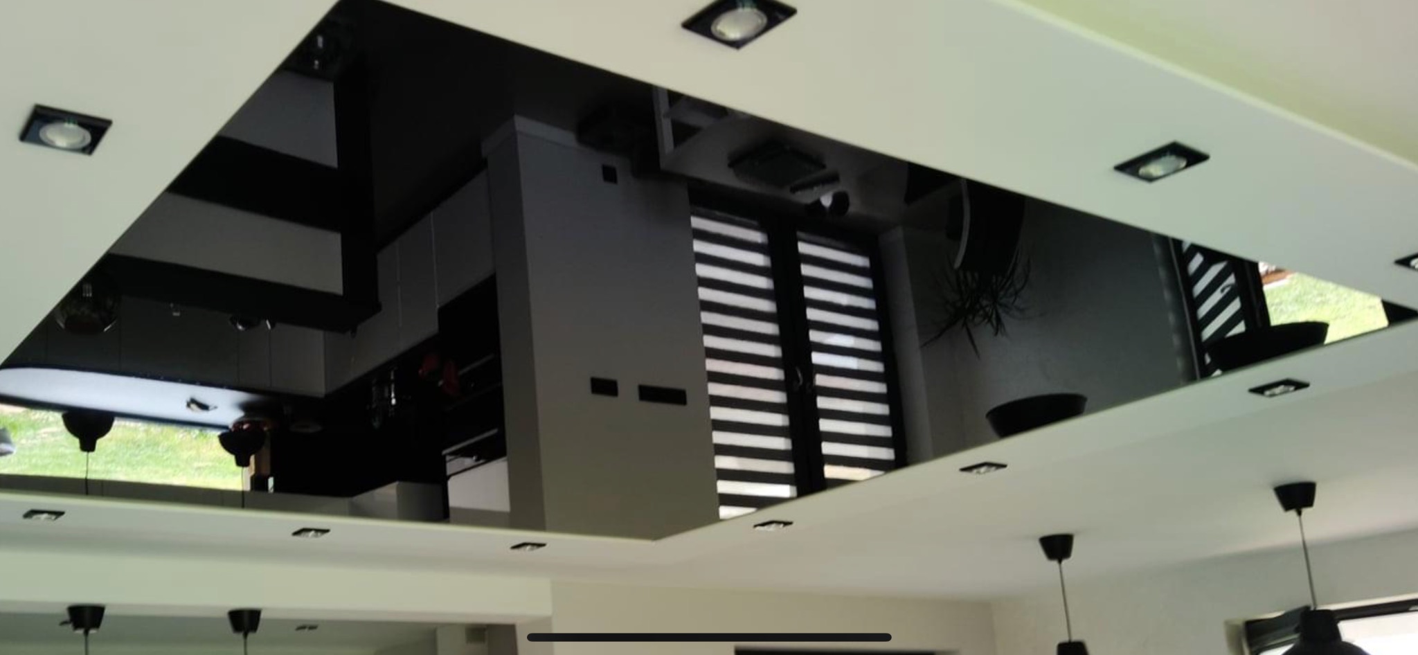 Błyszczący, czarny sufit lustrzany z wbudowanymi reflektorami, odbijający fragment nowoczesnej kuchni z szarymi szafkami i żaluzjami okiennymi.