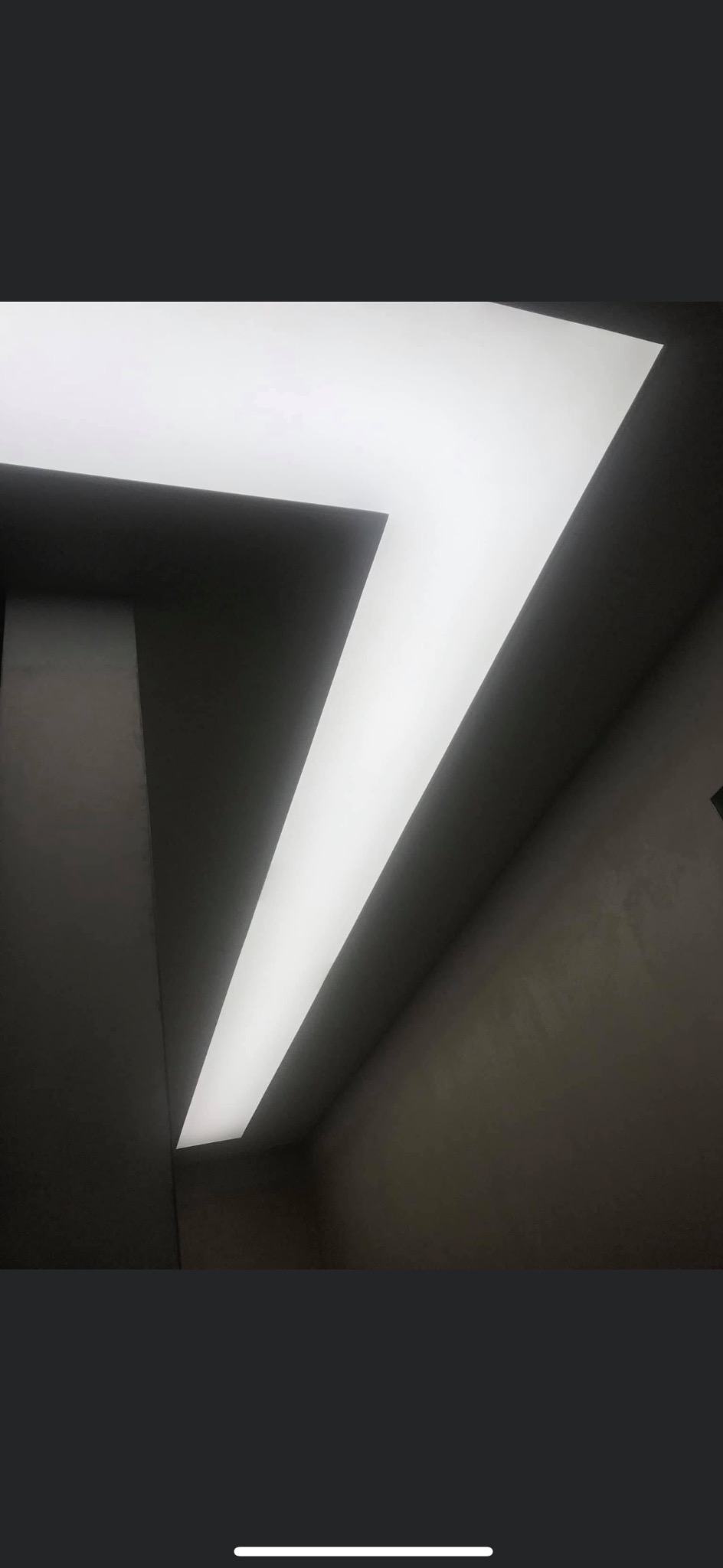 Geometryczna, podświetlana konstrukcja sufitowa w kształcie litery L, z ciemnymi ścianami w tle i delikatną teksturą, oświetlona równomiernym światłem.