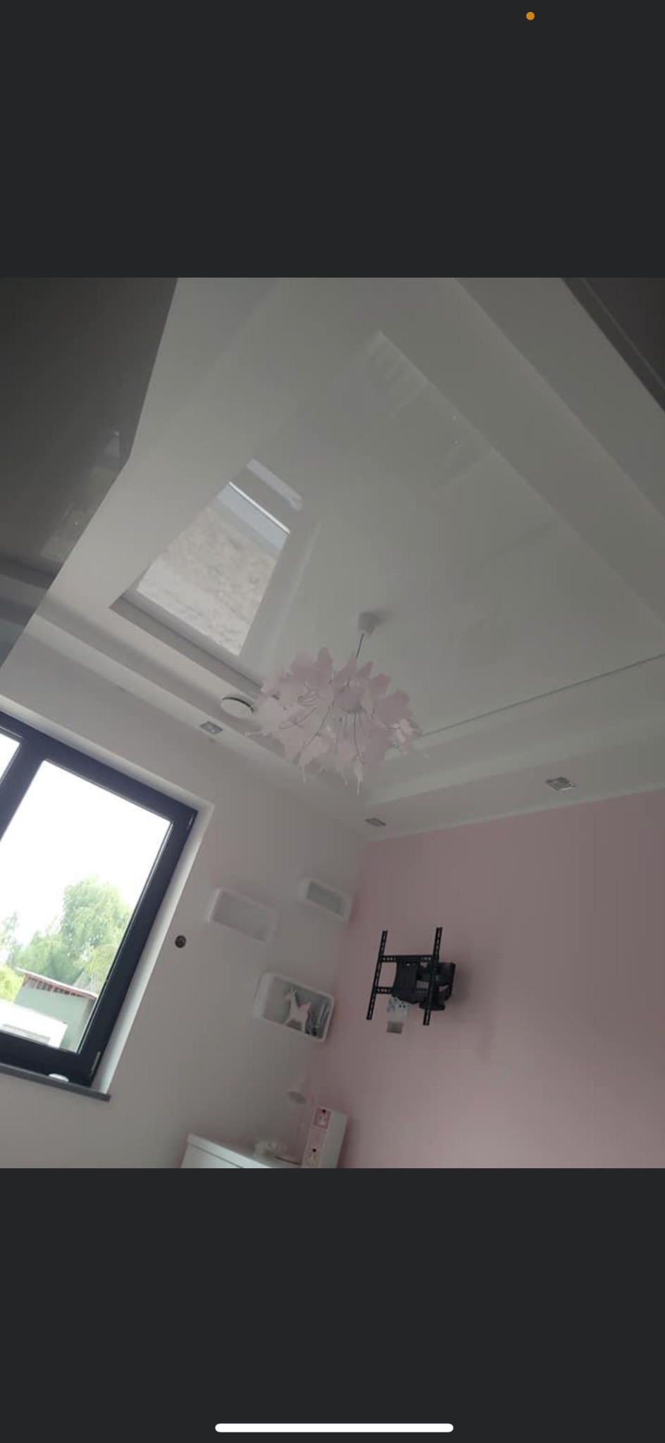 Błyszczący, biały sufit napinany z oknem dachowym i dekoracyjną, różową lampą w pokoju z różową ścianą i zamontowanym uchwytem na telewizor.