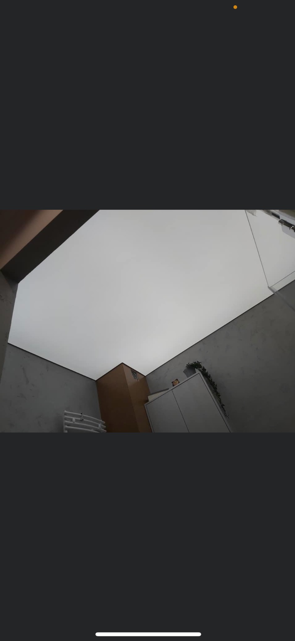 Biały sufit napinany w łazience z szarymi ścianami o teksturze betonu, widoczny grzejnik drabinkowy, szafka z rośliną i brązowy fragment ściany, ujęcie z dołu.