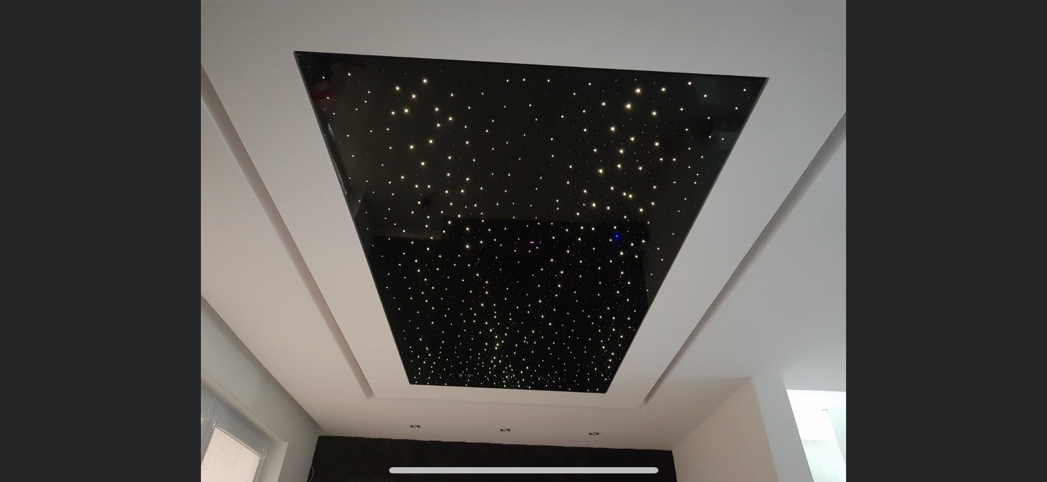 Czarny, trapezoidalny sufit podwieszany z efektem rozgwieżdżonego nieba, otoczony białymi listwami, widok od dołu.