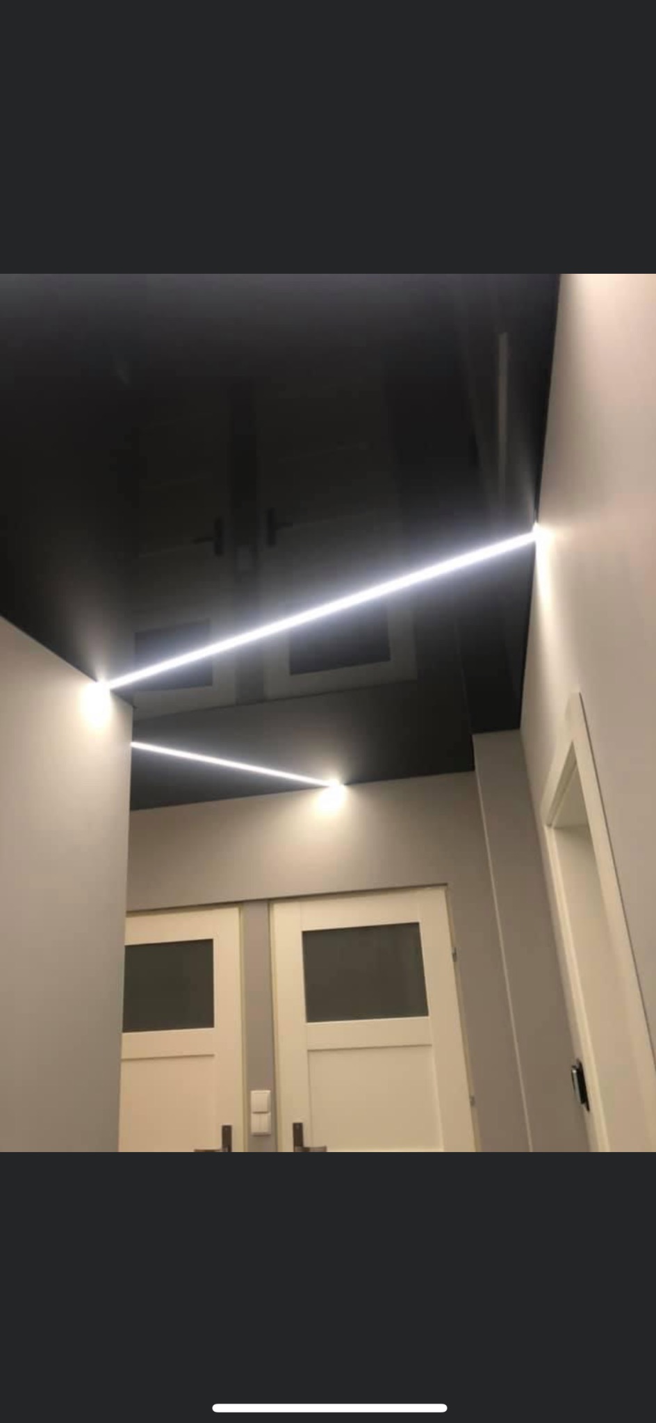 Czarne lustrzane wykończenie sufitu z wpuszczonymi liniowymi lampami LED, oświetlającymi szare ściany i białe drzwi z przeszkleniami.