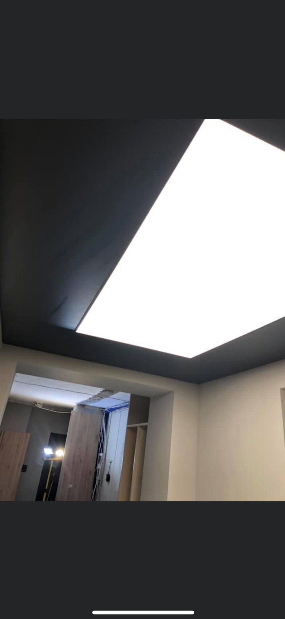 Pokój w trakcie prac wykończeniowych, widoczny sufit z zamontowanym panelem LED i czarną ramą, w tle drzwi z widocznym oświetleniem budowlanym.