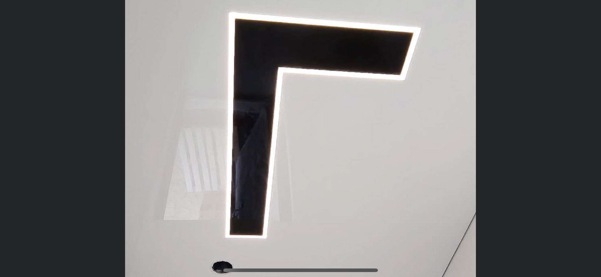 Czarny, geometryczny kształt litery L z podświetleniem LED na białej ścianie, widoczny otwór instalacyjny w dolnej części.