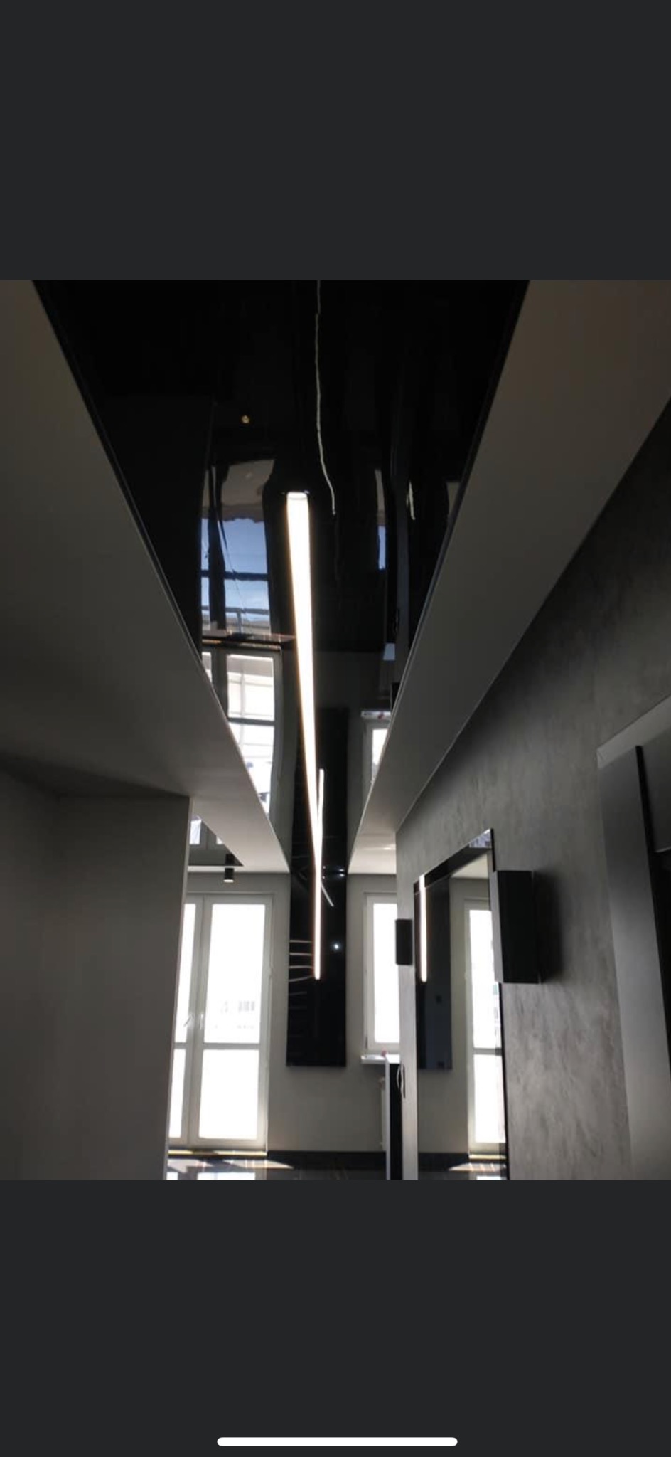 Nowoczesny korytarz z czarnym, lustrzanym sufitem podwieszanym i liniowym oświetleniem LED, szare ściany z efektem betonu, widoczne okna w głębi.